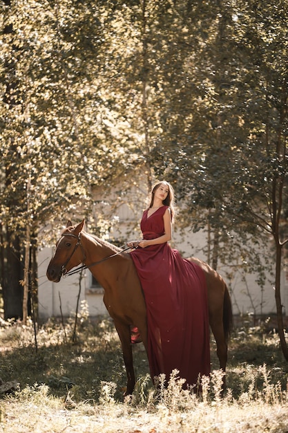 A beautiful young woman in a silk dress sits astride a horse. Horse ride on a sunny day in the forest.