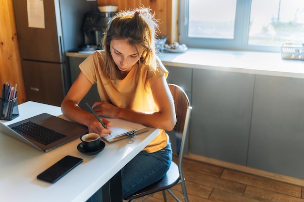 Beautiful young woman sit at desk studying or working on modern laptop at home in the kitchen