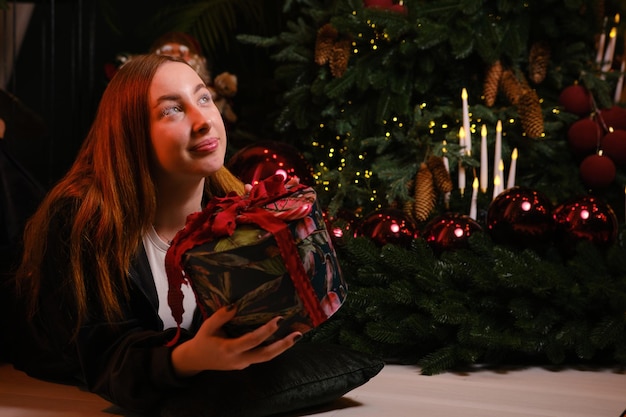 写真 美しい若い女性が黒い服を着て白い眉毛でクリスマスツリーの隣に座り赤いストライプと黒い背景の弓でプレゼントを握っています