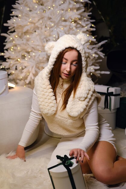 beautiful young woman sit in white dress and hat with white eyelashes next to christmas tree holding presents with green strip on dark background