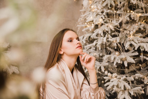 Beautiful young woman sits on a bed in an evening dress against the background of a Christmas tree