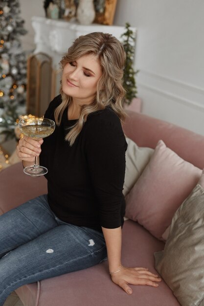 A beautiful young woman sits on the sofa with a glass of champagne near the christmas tree