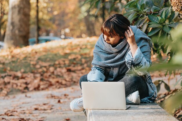 Beautiful young woman sitting on the bench in a park and working on a laptop while having fun taking