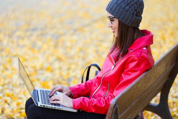 Foto bella giovane donna che si siede in una panchina e usando il suo computer portatile in autunno.