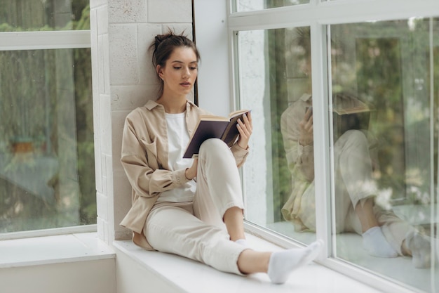 Beautiful young woman sitting by the window and reading a book