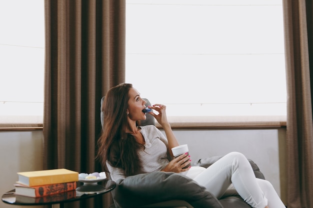 The beautiful young woman sitting on modern chair in front of window at home, eating macaroons and drinking coffee or tea