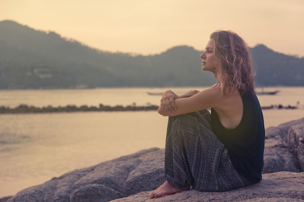 Bella giovane donna che si siede sulle rocce in riva al mare e guardando il tramonto