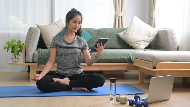 Beautiful young woman sitting in yoga mat and watching fitness lessons online on digital tablet