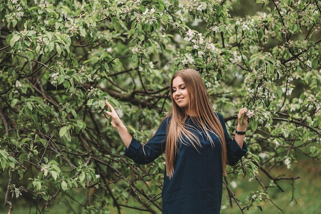 長いブロンドの髪を持つスラブの外観の美しい若い女性は、花の咲く緑の木の近くに立っています彼女は青いドレスを着ています