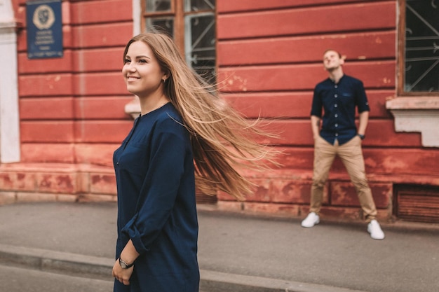 A beautiful young woman of Slavic appearance with long blond hair they develop in the wind She is wearing a blue dress A man is standing in the background selective focus