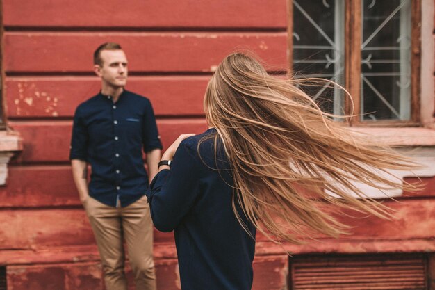 A beautiful young woman of Slavic appearance with long blond hair they develop in the wind She is wearing a blue dress A man is standing in the background selective focus