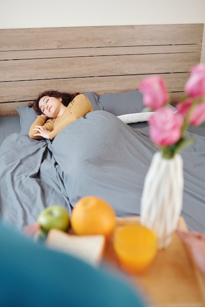 Beautiful young woman sleeping in bed when husband bringing her tray wih breakfast and flowers