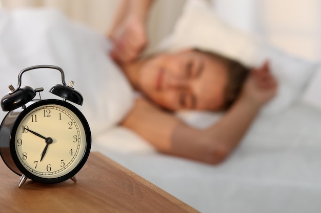 Beautiful young woman sleeping and smiling while lying in bed comfortably and blissfully on the background of alarm clock is going to ring Sunbeam dawn on her face