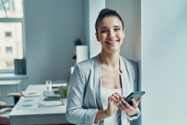 Beautiful young woman in smart casual using smart phone and smiling