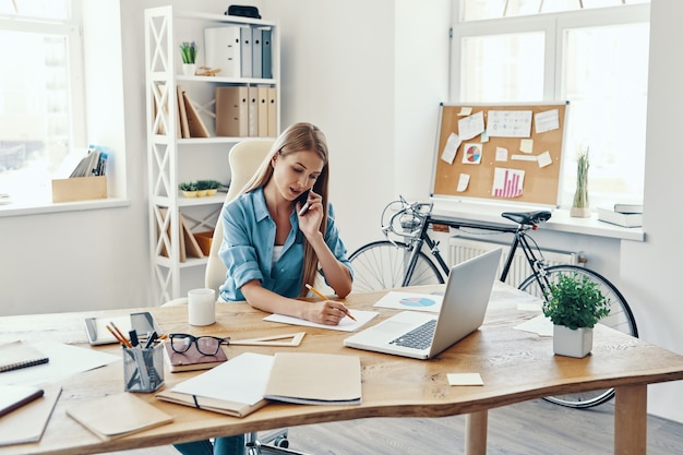 Beautiful young woman in smart casual wear writing something down and talking on the phone while sitting in the office