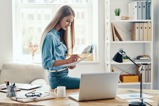 Beautiful young woman in smart casual wear writing something down and using laptop while sitting in the office