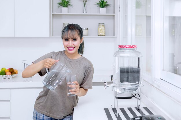 Photo beautiful young woman smiling at the camera while pouring fresh water into the glass in the kitchen room at home