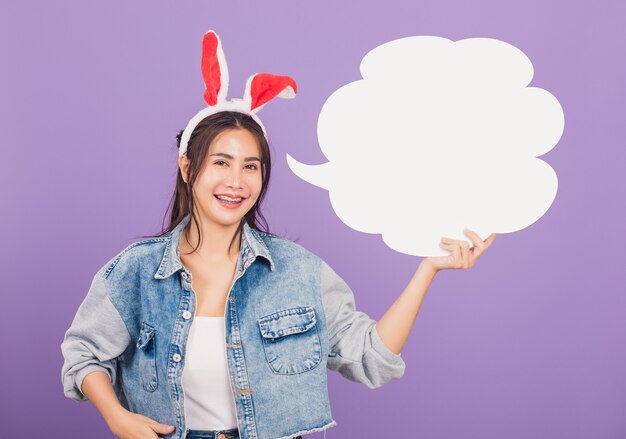 Beautiful young woman smiling excited wearing rabbit ears and denims holding empty speech bubble