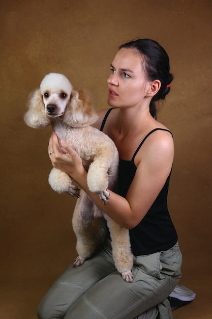 Beautiful young woman smiling and holding gorgeous dwarf white poodle dog. She sitting against brown studio wall and looking at dog.