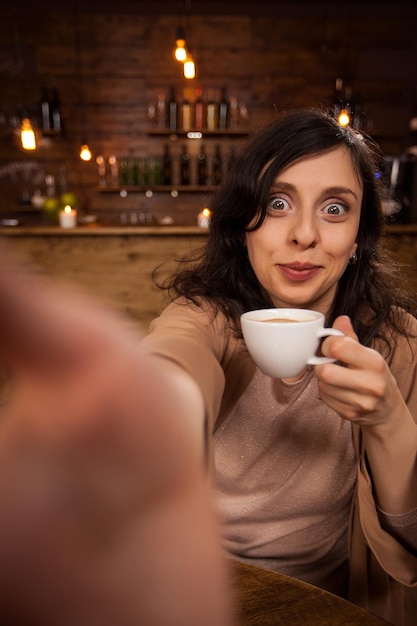 Красивая молодая женщина улыбается и с помощью смартфона делает автопортрет в кафе. Девушка развлекается со своим телефоном в кафе.