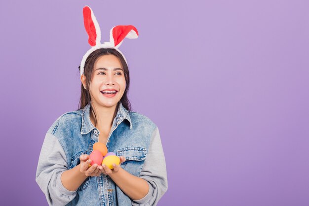 Красивая молодая женщина, улыбаясь в кроличьих ушах и джинсовой ткани, держит на руках красочный подарок пасхальных яиц