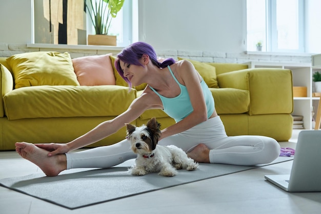 Beautiful young woman in sports clothing doing stretching exercises with her dog at home