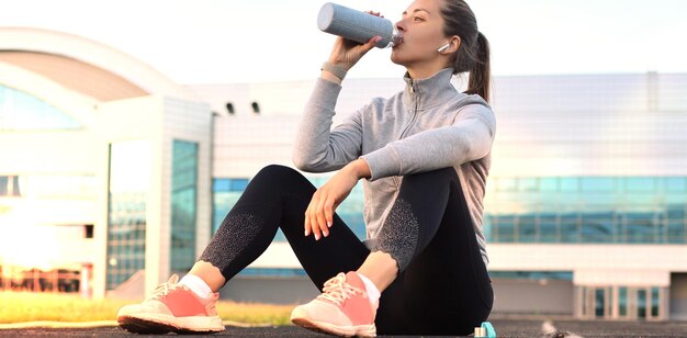 Красивая молодая женщина в спортивной одежде пьет воду после спортивных упражнений на открытом воздухе на стадионе.