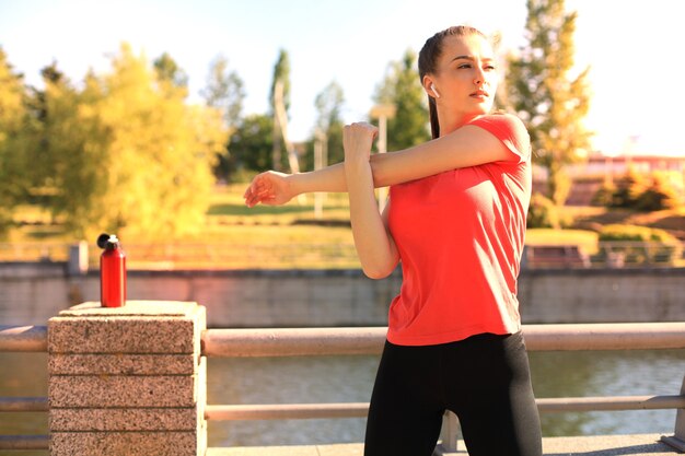 Beautiful young woman in sports clothing stretching her arms and looking concentrated while standing on the bridge.