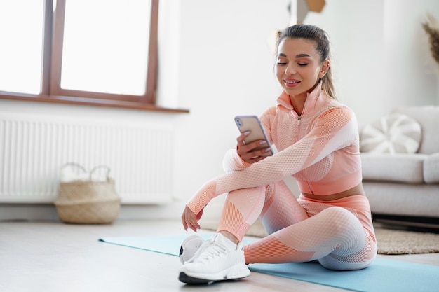 Beautiful young woman in sportswear sitting indoors and looking at smartphone before workout