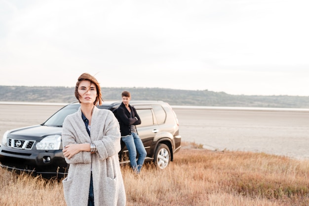 Beautiful young woman standing outdise with man leaning on car on the background