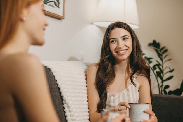 熱いお茶を飲みながらソファに座って彼女のガールフレンドと物語を語る美しい若い女性。