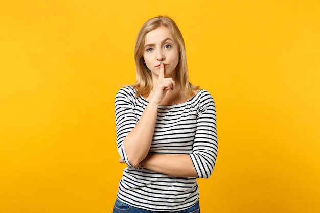 Beautiful young woman in striped clothes saying hush be quiet with finger on lips shhh gesture isolated on yellow orange wall background. People sincere emotions lifestyle concept. Mock up copy space.