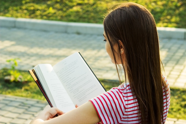 Beautiful young woman in striped t-shirt reading a book on the bench in the park. 