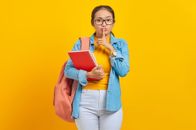 Beautiful young woman student in denim clothes with backpack and holding book fingers covering lips making silent gesture on yellow background education in high school university college concept