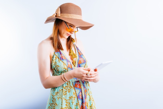 Bella giovane donna in abito estivo, cappello e occhiali gialli su sfondo bianco con copia spazio controllando il tablet dal lavoro. concetto di lavoro in vacanza