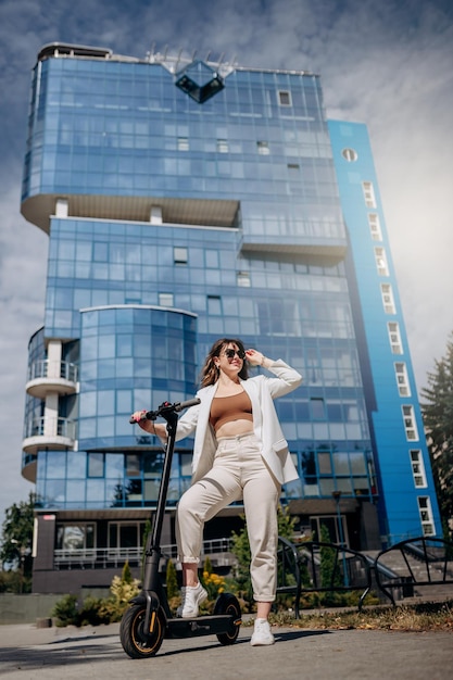 サングラスと白いスーツを着た美しい若い女性が、近代的な建物の近くの電動スクーターに立ち、目をそらす