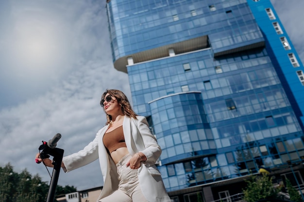 Beautiful young woman in sunglasses and white suit standing on her electric scooter near modern building and looking away