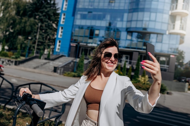 Bella giovane donna in occhiali da sole e abito bianco in piedi sul suo scooter elettrico vicino a un edificio moderno e fare selfie