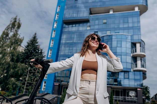 Beautiful young woman in sunglasses and white suit standing on her electric scooter near modern building and talking phone