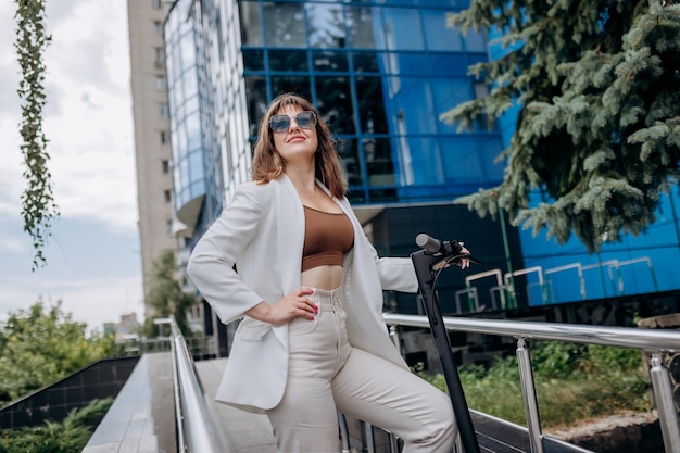 Bella giovane donna in occhiali da sole e abito bianco in piedi con il suo scooter elettrico vicino a un edificio moderno e distogliendo lo sguardo