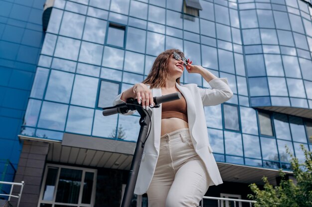 Beautiful young woman in sunglasses and white suit standing with her electric scooter near modern building and looking away