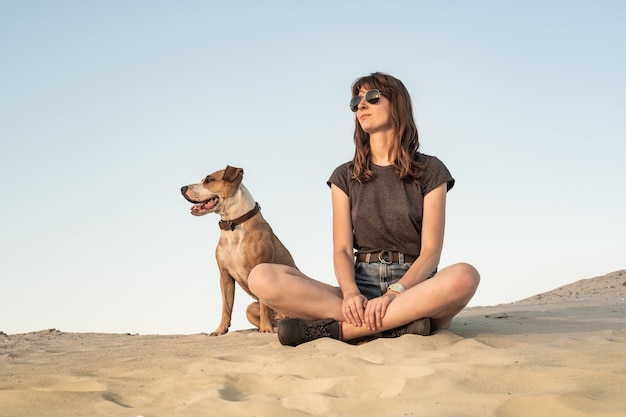 Beautiful young woman in sunglasses with dog sit on sand. Girl in hiking casual clothes and staffordshire terrier puppy sitting on sandy beach or in desert on hot sunny day