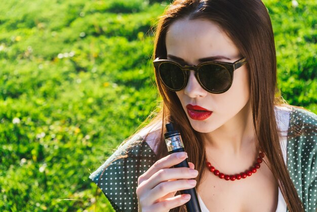 Beautiful young woman in sunglasses with long hair sitting on lawn with bright green grass, smoking. sunny day