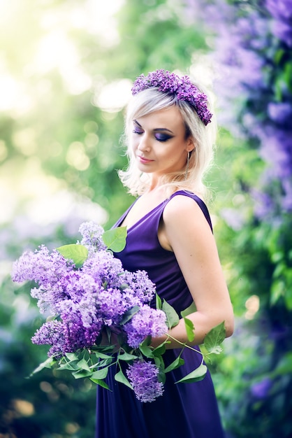 Красивая молодая женщина, в окружении фиолетовых цветов. женщина сидит в длинном платье с разрезом на фоне весеннего сада с сиренью