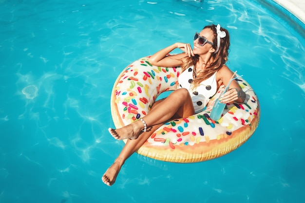 Beautiful young woman in swimming pool swims on inflatable ring donut and has fun with glass of cocktail on vacation.