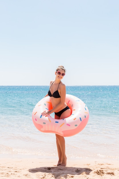 Beautiful young woman in swimming pool swims on inflatable ring donut and has fun with glass of cocktail on vacation.