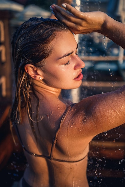 Красивая молодая женщина в купальнике с идеальной фигурой в душе в лучах заката у моря Девушка с золотым загаром в отпуске