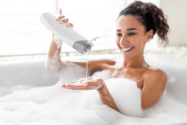 Foto bella giovane donna che prende il bagno di bolle versando lo shampoo in mano godendosi la procedura della spa domestica