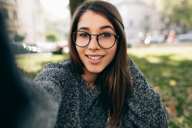 スマートフォンで自画像を撮る美しい若い女性女性は街の通りで自分撮りを作るニットセーター透明な眼鏡を着ています
