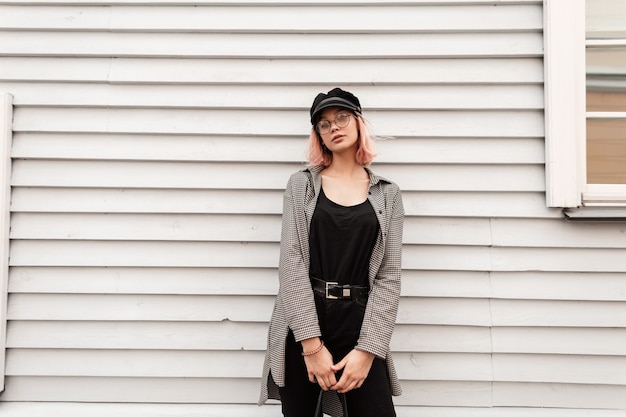 Beautiful young woman teenager in fashion casual clothes with vintage glasses and a hat stands near a wooden house wall